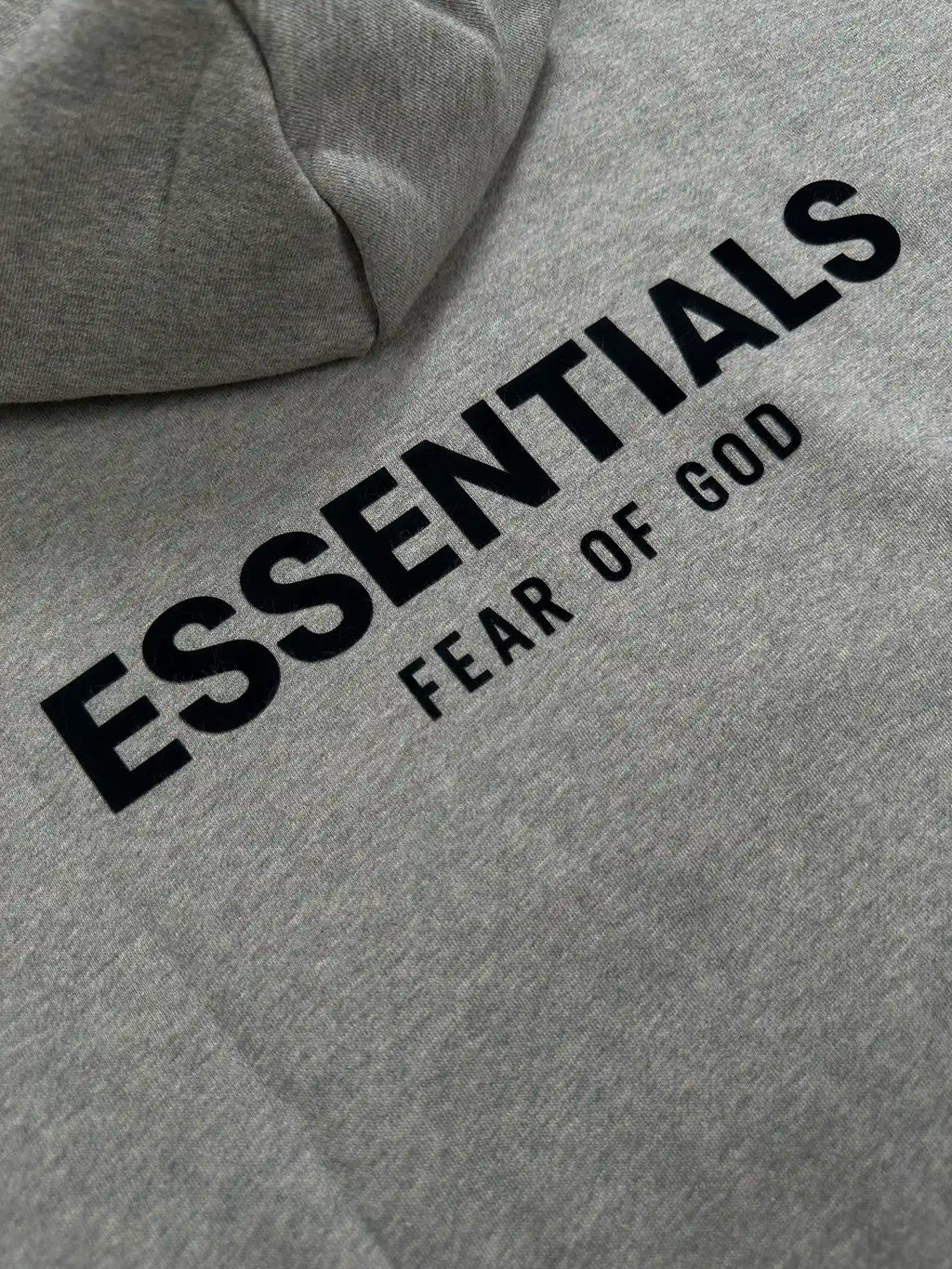 Close-up of the back logo detail on a light heather grey ESSENTIALS hoodie, featuring raised black lettering on soft cotton blend fabric with a relaxed streetwear fit.