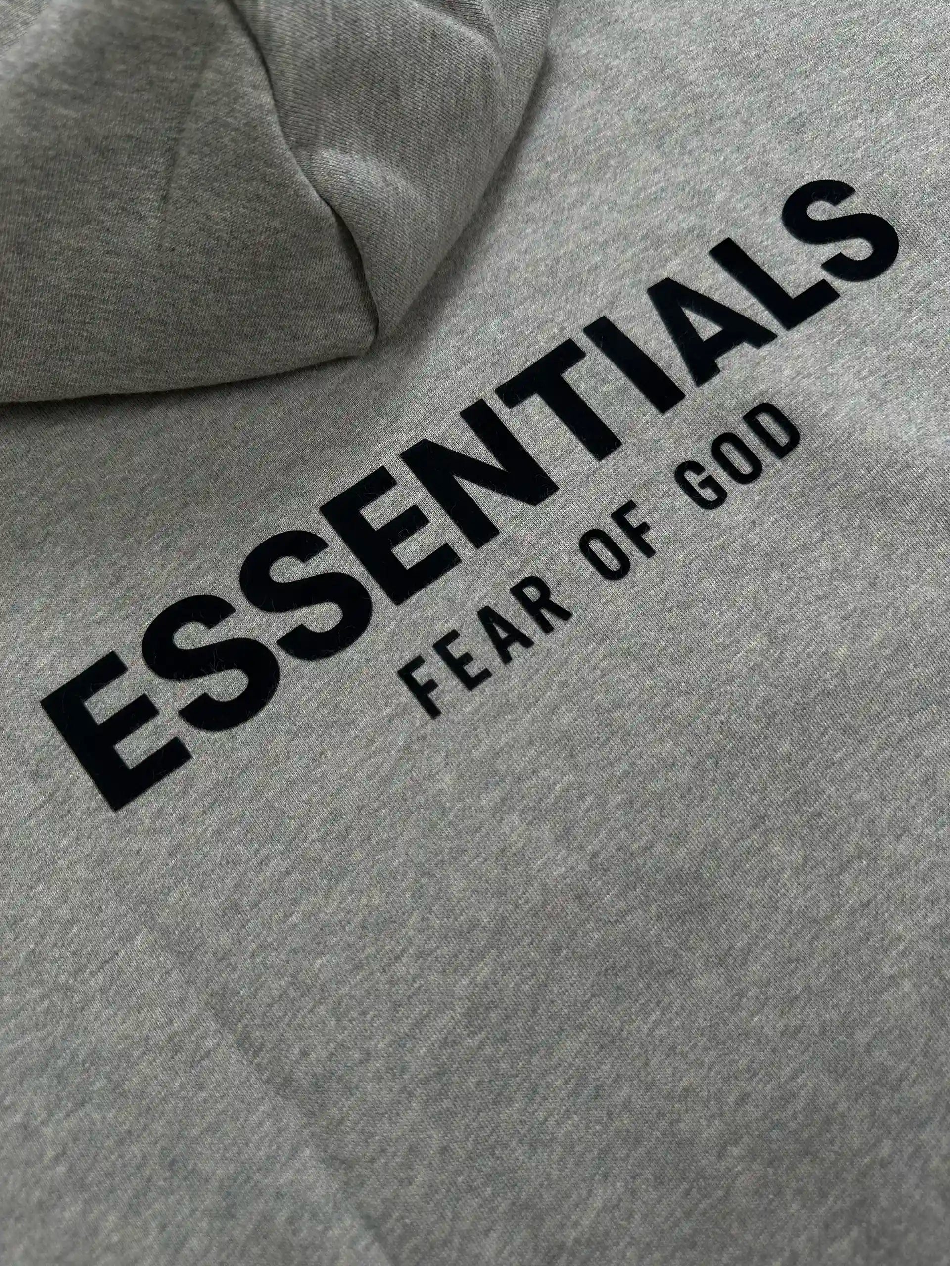 Close-up of the back logo detail on a light heather grey ESSENTIALS hoodie, featuring raised black lettering on soft cotton blend fabric with a relaxed streetwear fit.