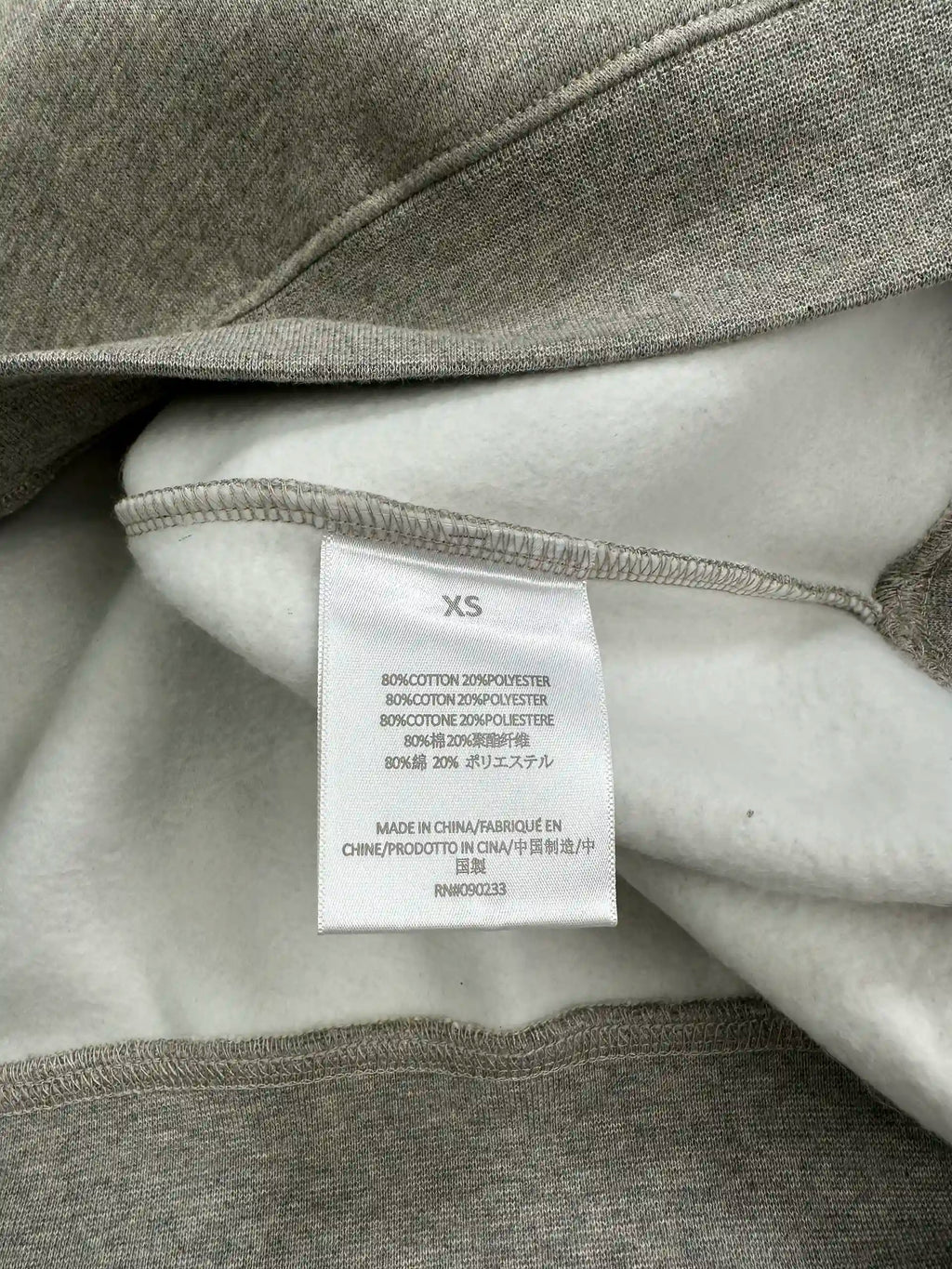 Fabric content and size label inside a light heather grey ESSENTIALS hoodie, showing cotton and polyester material composition, interior fleece texture, and manufacturing origin details.