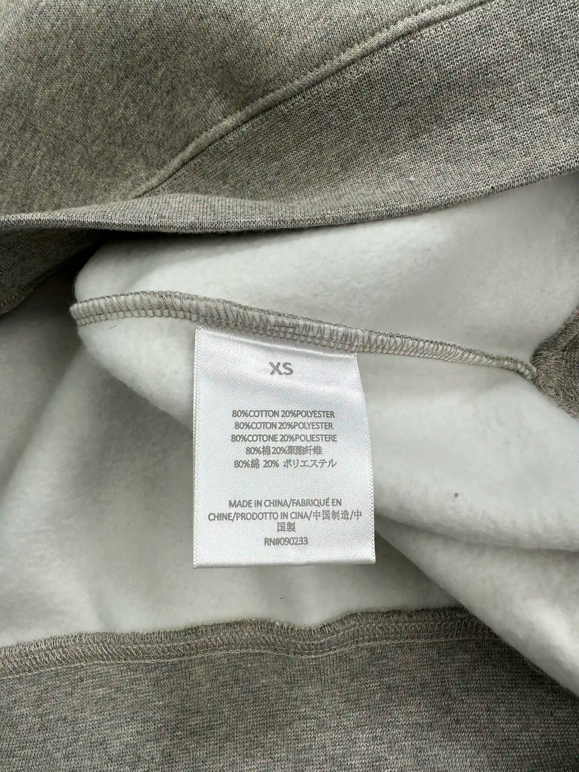 Fabric content and size label inside a light heather grey ESSENTIALS hoodie, showing cotton and polyester material composition, interior fleece texture, and manufacturing origin details.