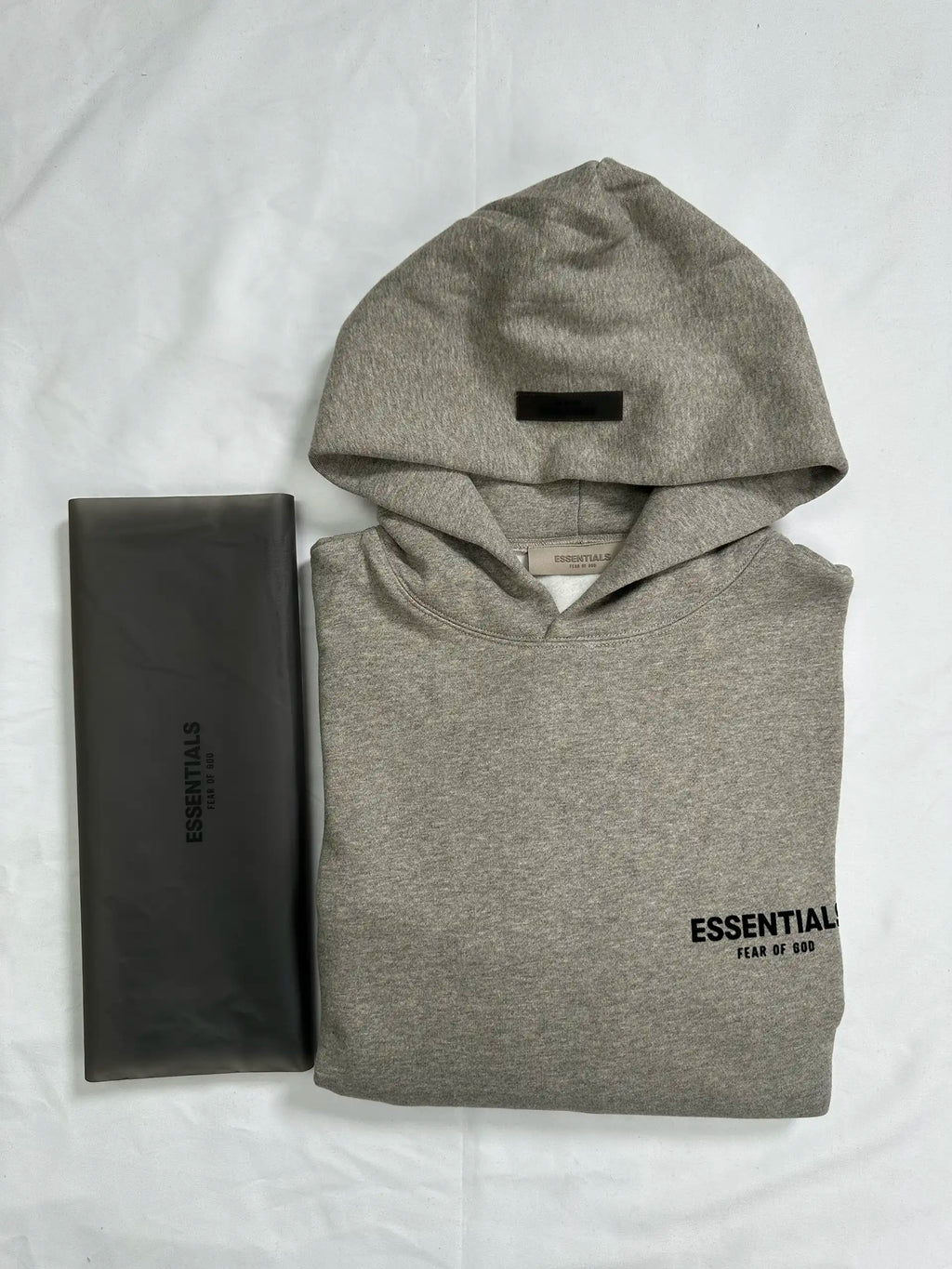 Folded light heather grey ESSENTIALS hoodie displayed flat with hood visible and original semi-transparent zip packaging placed beside it, showcasing minimalist streetwear design, clean construction, and retail presentation.
