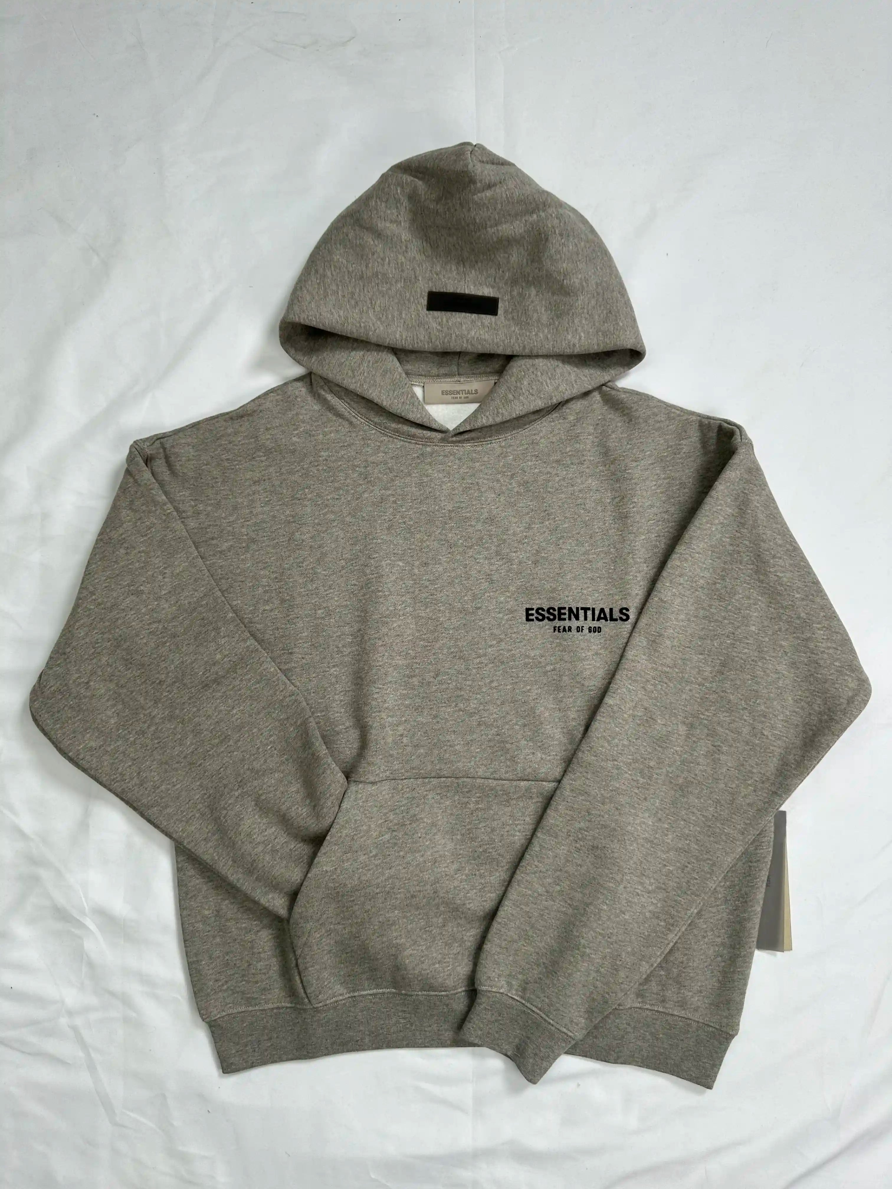 Front view of a light heather grey ESSENTIALS hoodie laid flat on a white background, featuring a relaxed oversized fit, ribbed cuffs and hem, kangaroo pocket, and small minimalist chest logo, showcasing clean streetwear design and everyday casual style.