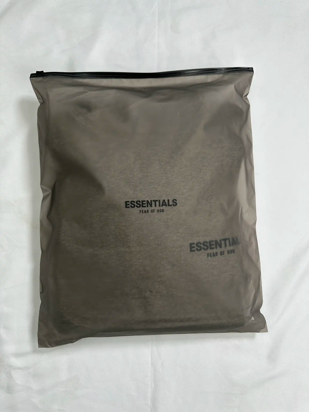Retail packaging view of a light heather grey ESSENTIALS hoodie sealed in a semi-transparent zip bag, showing the folded garment and minimalist logo placement through the packaging.