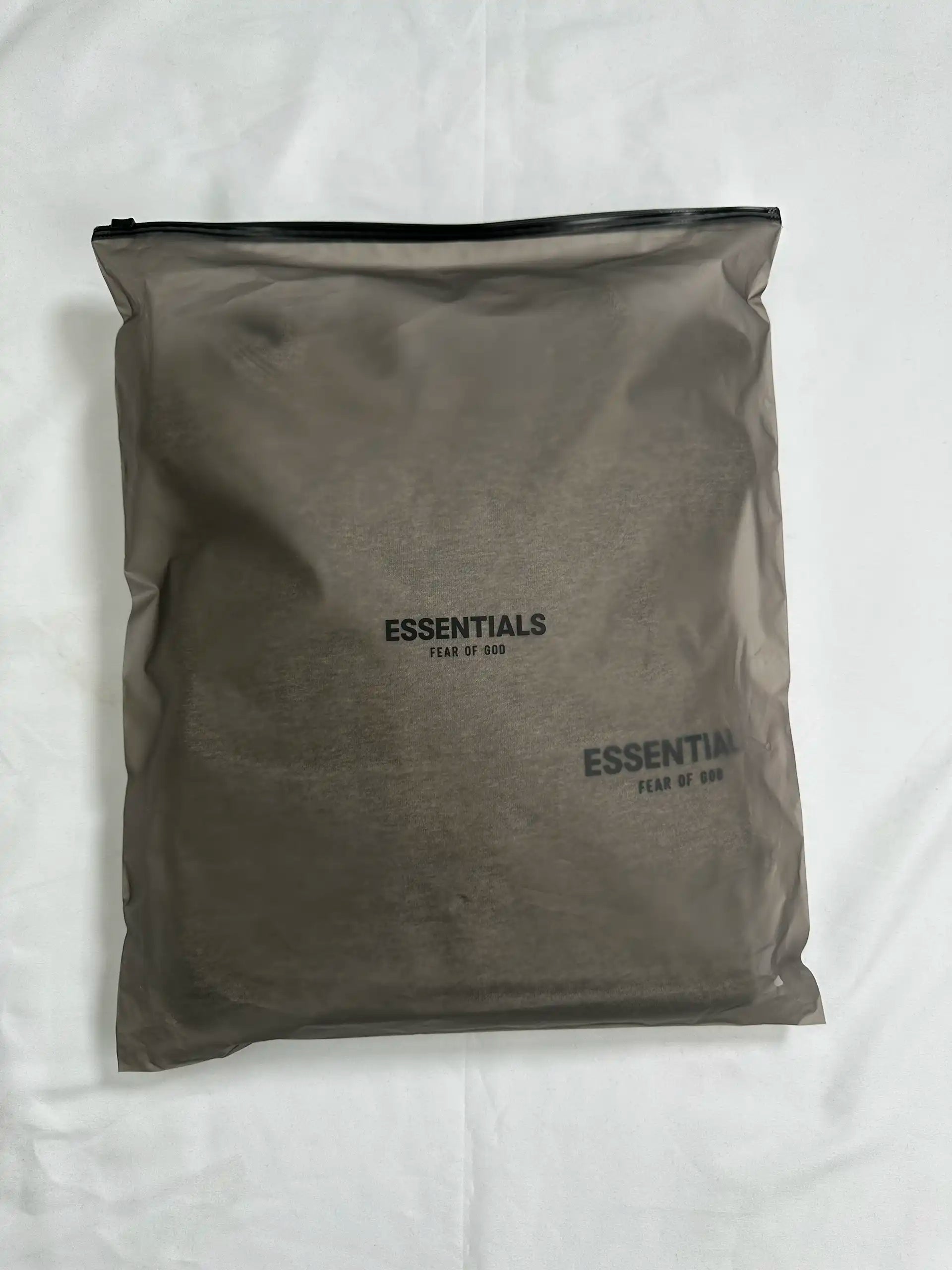 Retail packaging view of a light heather grey ESSENTIALS hoodie sealed in a semi-transparent zip bag, showing the folded garment and minimalist logo placement through the packaging.