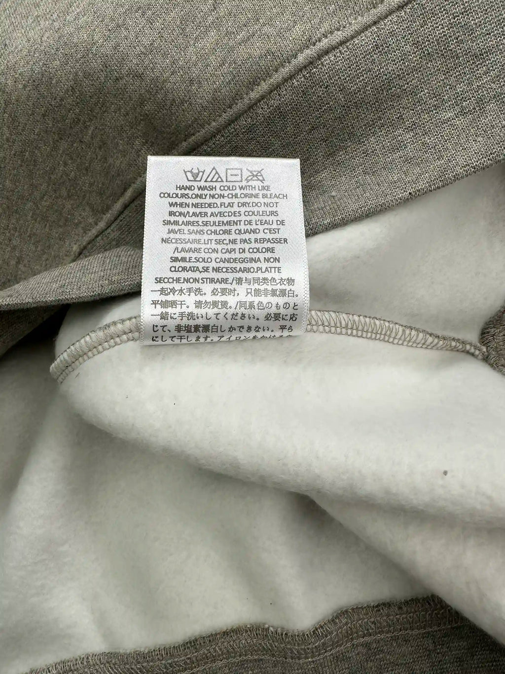 Wash care instruction label inside a light heather grey ESSENTIALS hoodie, displaying garment care symbols, washing guidelines, and multilingual care instructions on the interior lining.